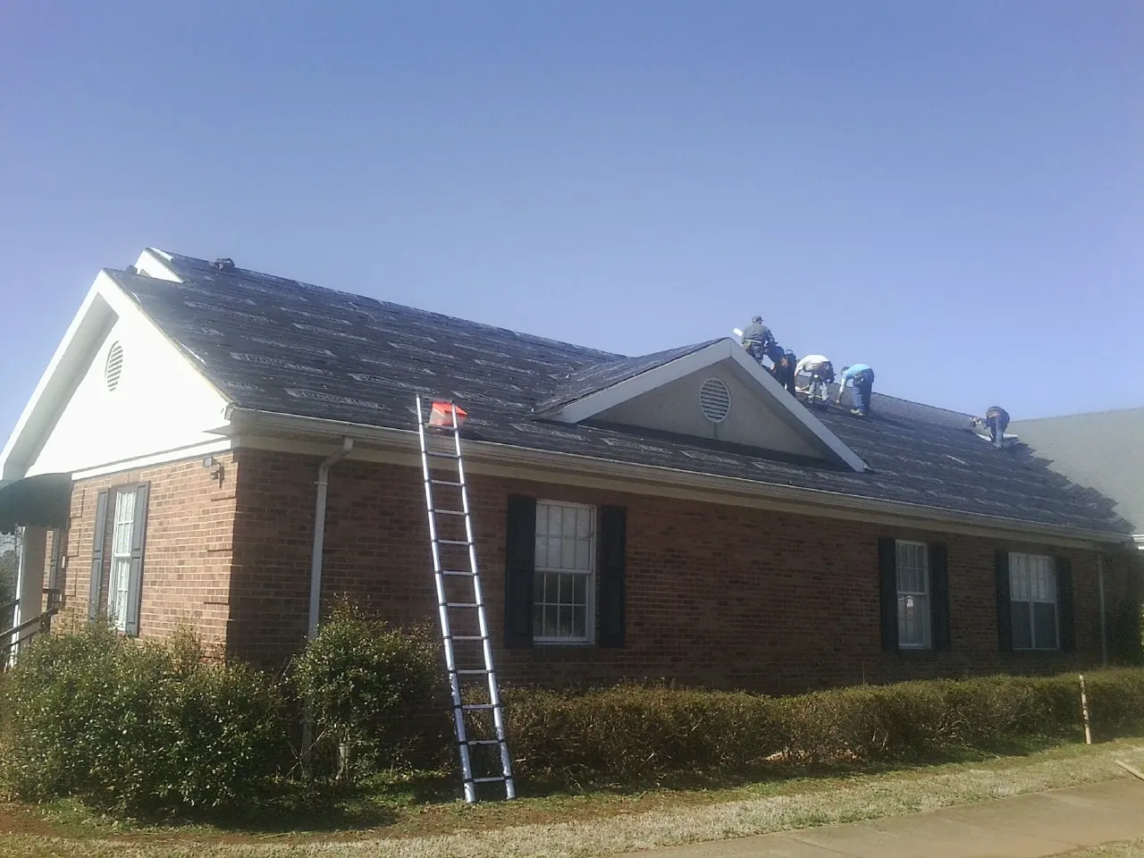 Crew tearing shingles off a brick ranch roof with a ladder set — Emergency Roof Repair work by our crew in Wayland